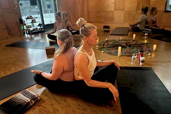 Two women sitting back-to-back on a yoga mat in a studio.